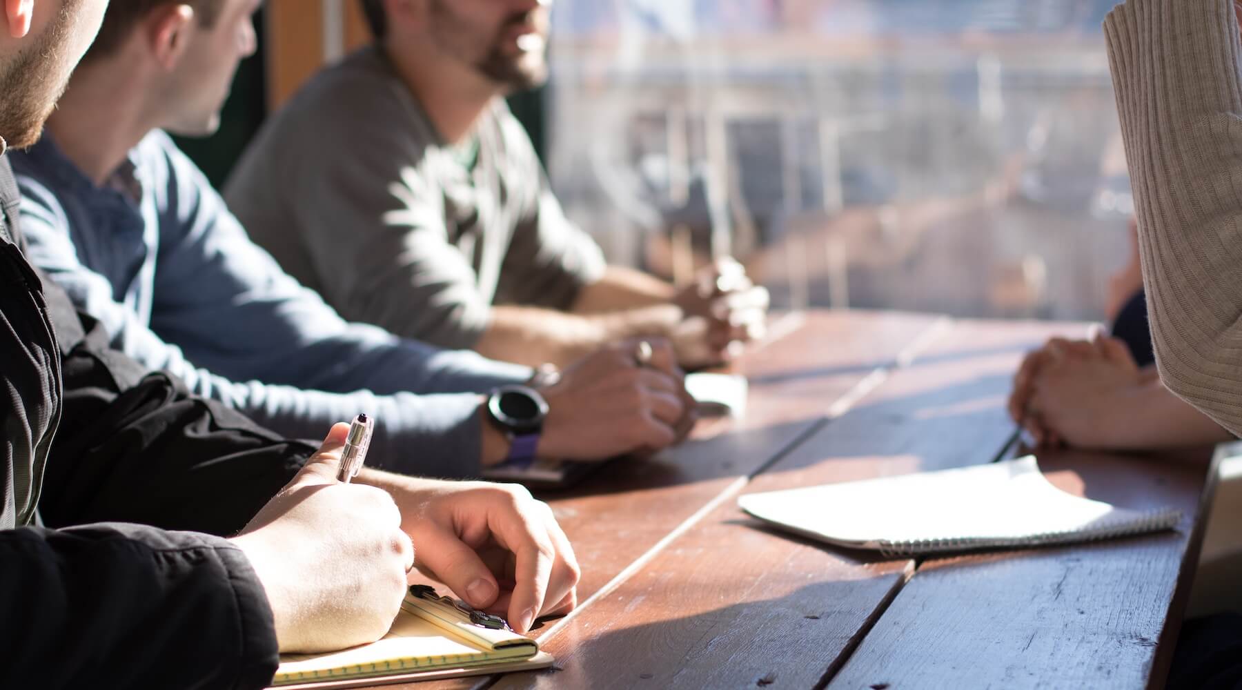 People taking notes during a business meeting around a wooden table.