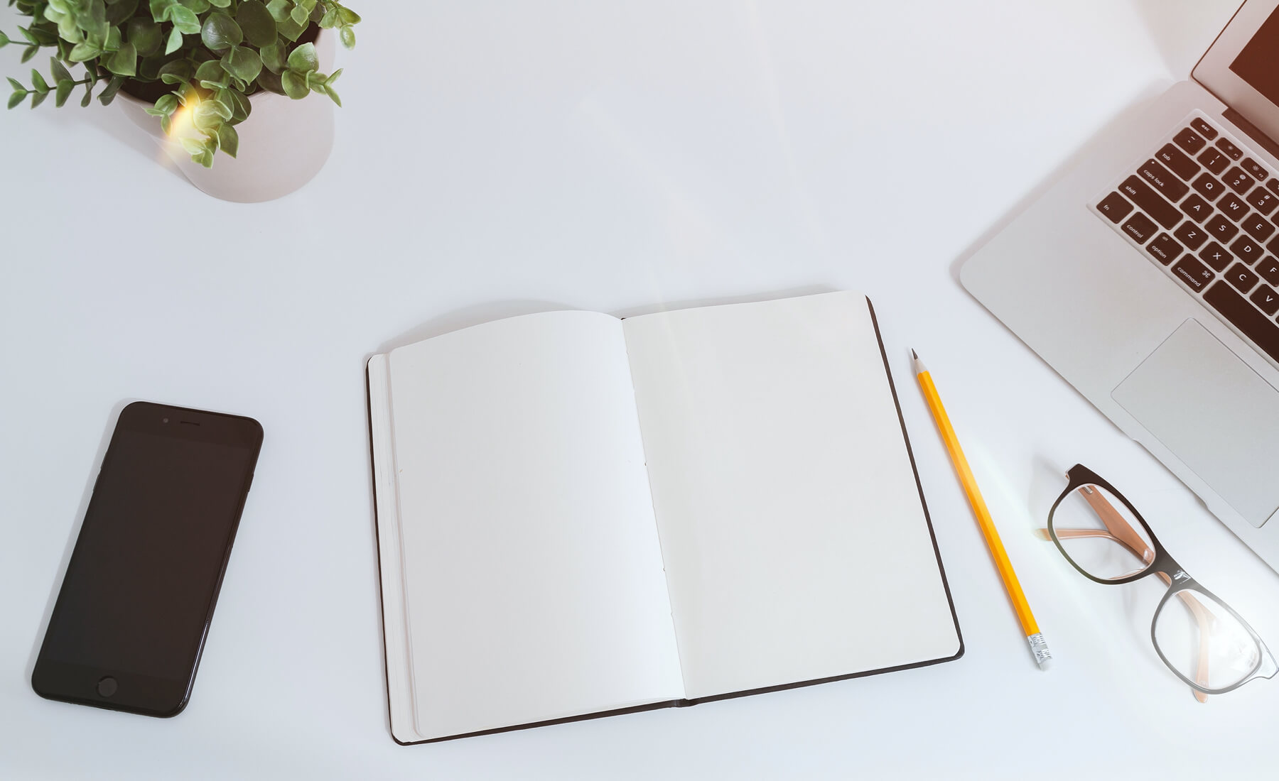 Flat-lay of an office desk with a phone, open notebook, glasses, pencil, and laptop.