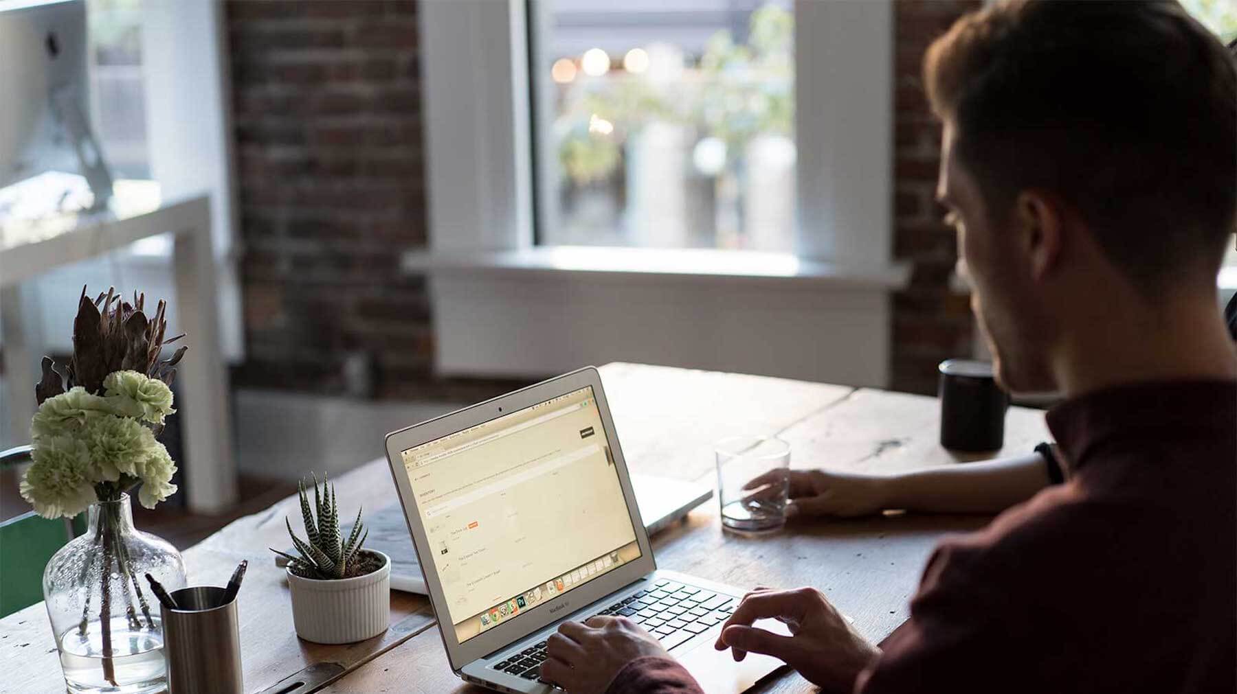 Person typing on a laptop at a wooden desk with flowers and plants nearby.