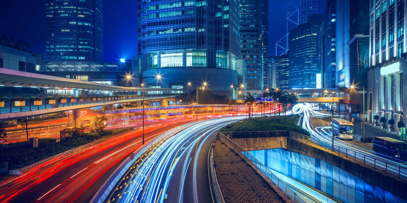 Nighttime cityscape with light trails from highway traffic and skyscrapers in the background.